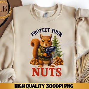 May include: Beige sweatshirt with the text "PROTECT YOUR NUTS" in navy and red. The graphic features a squirrel in a camouflage jacket holding a gun, with a small pine tree and acorns. The words "HIGH QUALITY 300DPI PNG" are at the bottom.