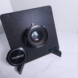 May include: A black Tamron lens attached to a black Toyo camera back. The lens has a silver ring around the edge and a red dot on the top.