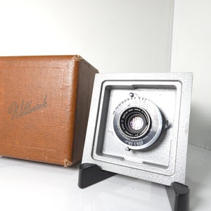 May include: A silver camera body with a black lens. The camera is sitting on a black stand. The camera is in a brown box with the word "Willard" printed on it.