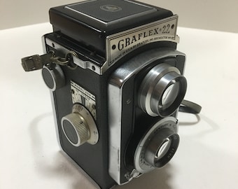 Może przedstawiać: Czarno-srebrny aparat Graflex 22 z paskiem skórzanym. Aparat ma srebrny obiektyw i czarne korpus z srebrną tabliczką znamionową z napisem "Graflex 22 Made in U.S.A. by Graflex Inc. Rochester NY".