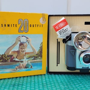 May include: A vintage Kodak Brownie Flashmite 20 camera outfit in its original box. The camera is silver and blue with a flash attachment. The box is yellow with a picture of a girl in a swimming pool.