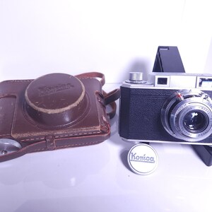 May include: A silver Konica rangefinder camera with a black leather case. The camera has a silver lens with black markings and a black body. The case is brown leather with a silver buckle.