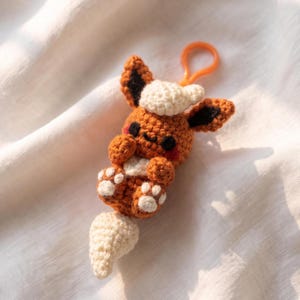 May include: A brown and cream-colored crochet keychain shaped like a small creature with pointed ears, black eyes, pink cheeks, and a fluffy tail. The figure features a cream-colored tuft on its head and paws with white accents, attached to an orange plastic ring.