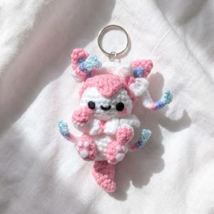 May include: A small, handcrafted crochet keychain featuring a creature with white and pink yarn, black embroidered eyes, and a smiling mouth. The figure has long, flowing ears and tail accents in pastel blue, pink, and white yarn, attached to a metal ring.