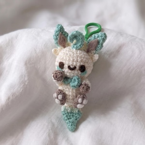May include: A small, handmade crochet plush keychain featuring a cream-colored creature with mint green accents on its ears, tail, and neck. It has dark brown embroidered eyes, a small smile, and brown paws with white detailing, attached to a green plastic clip.