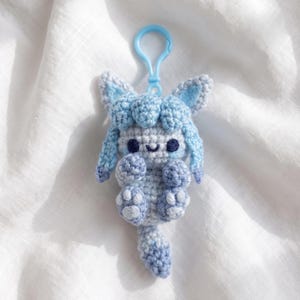 May include: A light blue and white crocheted creature keychain featuring a small animal-like figure with pointed ears, a rounded head, and a long tail. The figure has dark blue eyes and a small stitched smile, attached to a light blue plastic clasp.