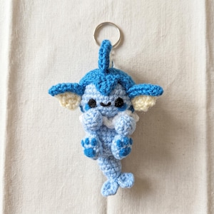 May include: A crochet keychain featuring a small, blue creature with a large head, pointed ears with cream-colored inner sections, a white collar, and a long tail. The figure has black bead eyes and a small stitched mouth, attached to a silver ring.