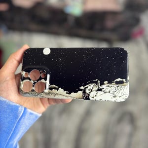 Op de afbeelding: Zwarte telefoonhoes met een wit ontwerp van een persoon die onder een sterrenhemel met een maan ligt.