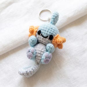 May include: A light blue crocheted axolotl keychain featuring orange external gills, a small tail, and embroidered black eyes and a mouth. The creature has light purple accents on its feet and is attached to a silver metal keyring loop.