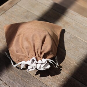 May include: A brown drawstring bag with a white ruffled trim at the opening. The bag is made of a soft, textured material and is gathered at the top with a white drawstring. The bag is sitting on a wooden surface.