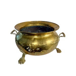 May include: A vintage brass cauldron with a wide rim and two handles. The cauldron rests on three claw feet. The brass has a golden hue with some areas showing signs of age and wear. The interior is dark.