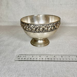 May include: A silver bowl with a raised rim decorated with a repeating pattern of pears and leaves. The bowl sits on a short, round pedestal.