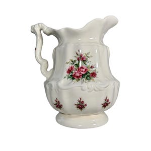 May include: A white ceramic pitcher with a floral rose design. The pitcher has a curved handle and a spout. The roses are pink and red with green leaves. The pitcher is approximately 10 inches tall.
