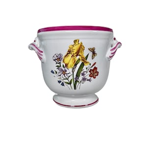 May include: White ceramic planter with a floral design featuring yellow irises, butterflies, and other flowers. The planter has a pink rim and handles, and a pedestal base. A decorative piece for home decor.