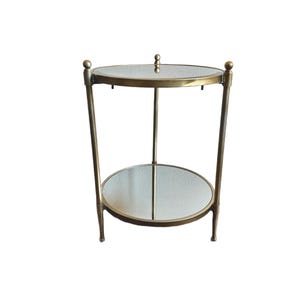 May include: A two-tiered side table with a gold-colored metal frame. The table features two circular glass shelves. The legs are slightly angled, and the top of the table has a small decorative finial. The table is approximately 24 inches tall.