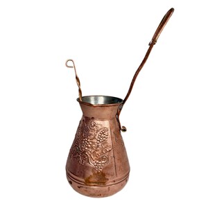 May include: A copper-colored Turkish coffee pot, also known as a cezve, with a long handle and a small, decorative spoon. The pot features an embossed grape vine design on its body. The cezve is designed for brewing coffee.