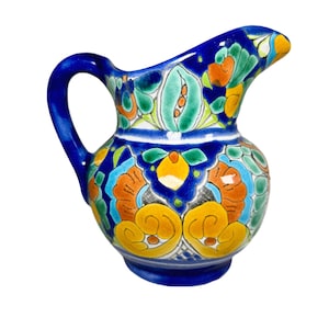 May include: A small, hand-painted ceramic pitcher with a vibrant floral design. The pitcher features a deep blue base and handle, with colorful accents of orange, yellow, green, and turquoise. The spout is gently curved, and the overall design is ornate.