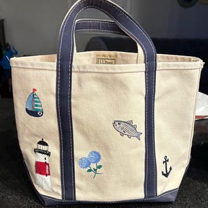 Puede incluir: Bolso tote de lona color crema con asas y ribetes azul marino. El bolso presenta diseños náuticos bordados, incluyendo un velero, un faro, un pez, flores azules y un ancla. La marca "L.L.Bean" es visible.