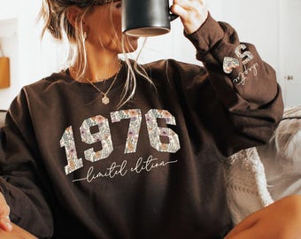 May include: A dark brown sweatshirt featuring the year "1976" in a floral pattern, with "limited edition" below. The sleeve has a floral heart and the word "chapter". A black mug is held up to the face.