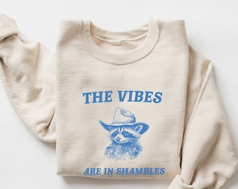 Peut inclure: Un sweat-shirt beige avec un graphique bleu d'un raton laveur portant un chapeau de cowboy. Le texte "THE VIBES ARE IN SHAMBLES" est imprimé en bleu sous le raton laveur.