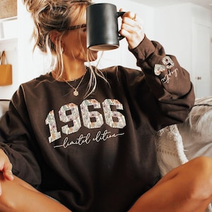 May include: A dark brown sweatshirt with the year "1966" in a floral design. The words "limited edition" are below the year. The sleeve has a small heart design with the number "59" and the word "chapter". A black mug is held up to the face.
