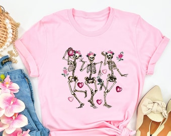 May include: Pink t-shirt with a graphic of three skeletons wearing floral crowns and holding hearts. The skeletons are dancing and surrounded by flowers.