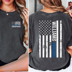 May include: Dark gray t-shirt featuring a distressed American flag design with a blue line, and the name "Williams" on the back. The front of the shirt has a small flag with a blue line. The shirt is made of soft material.