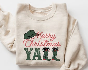 Può includere: Una felpa beige con il testo "Merry Christmas Y'all" in rosso e verde. Un cappello da cowboy e due paia di stivali con bastoncini di zucchero sono anche sulla felpa.