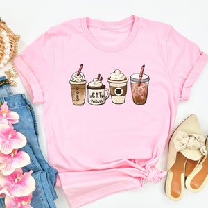 以下が含まれることがあります： コーヒーの飲み物が4種類描かれたピンクのTシャツ。飲み物はキャラメルマキアート、ラテ、モカ、ストロベリーリフレッシュメント。モカのカップには「Cat Mom」の文字が書かれている。
