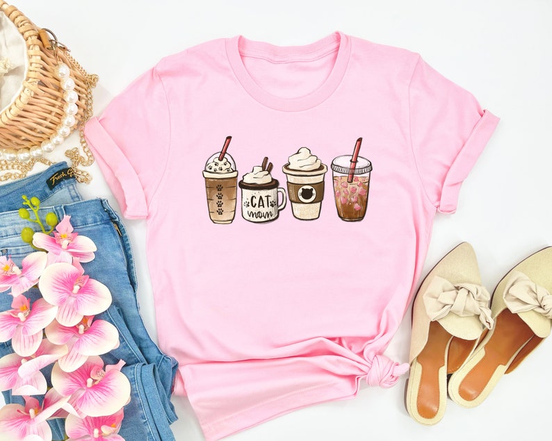 以下が含まれることがあります： コーヒーの飲み物が4種類描かれたピンクのTシャツ。飲み物はキャラメルマキアート、ラテ、モカ、ストロベリーリフレッシュメント。モカのカップには「Cat Mom」の文字が書かれている。