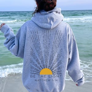 May include: A gray hoodie with a black sunburst design and the text "BE THE LIGHT" and "MATTHEW 5:14" on the back. The person wearing the hoodie is standing on a beach.