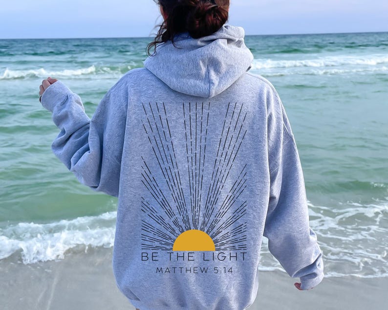 May include: A gray hoodie with a black sunburst design and the text "BE THE LIGHT" and "MATTHEW 5:14" on the back. The person wearing the hoodie is standing on a beach.