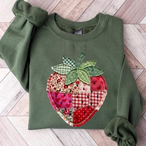 May include: An olive green crewneck sweatshirt featuring a large, central strawberry-shaped appliqu&eacute;. The strawberry is constructed from various fabric patches in shades of red, pink, and white, featuring floral, heart, and checkered patterns.