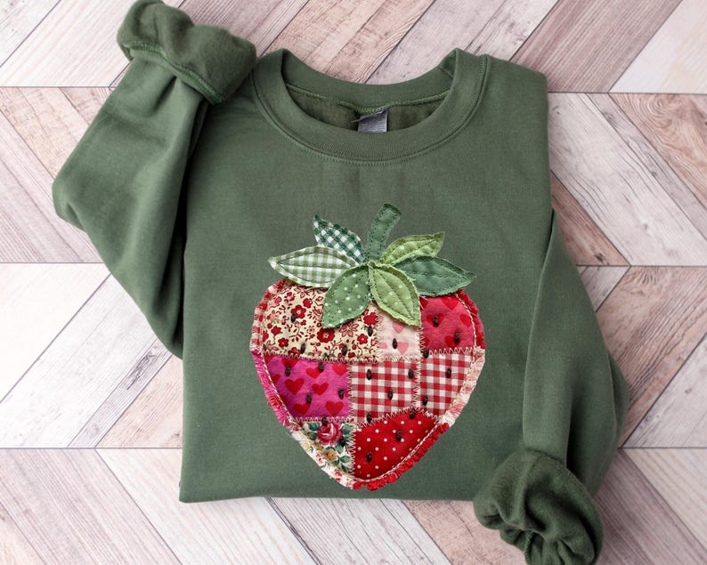 May include: An olive green crewneck sweatshirt featuring a large, central strawberry-shaped appliqu&eacute;. The strawberry is constructed from various fabric patches in shades of red, pink, and white, featuring floral, heart, and checkered patterns.
