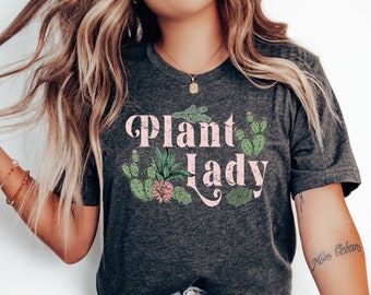 Könnte beinhalten: Ein dunkelgraues T-Shirt mit dem Text "Plant Lady" in rosa Schrift. Der Text ist von einer Grafik mit verschiedenen grünen und rosa Kakteen und Sukkulenten umgeben.