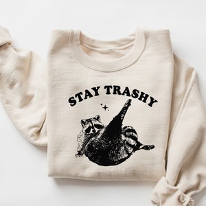 Op de afbeelding: Een beige sweatshirt met een zwarte afbeelding van een wasbeer met de tekst "STAY TRASHY" erop gedrukt.