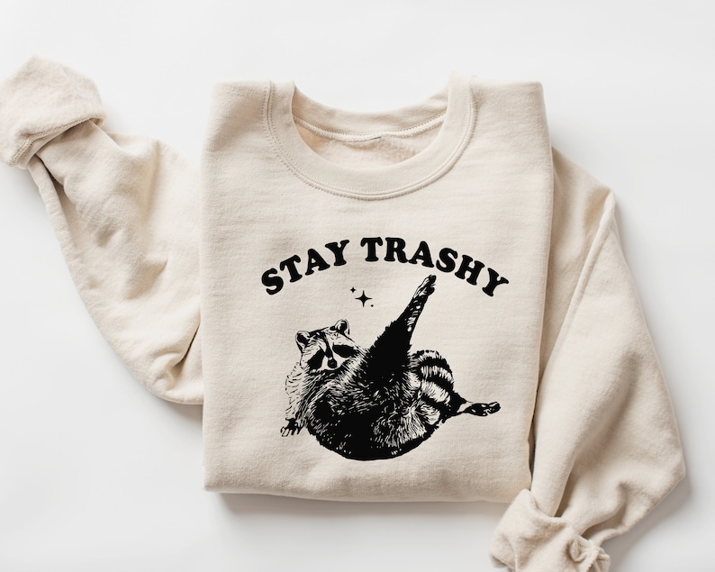 Op de afbeelding: Een beige sweatshirt met een zwarte afbeelding van een wasbeer met de tekst "STAY TRASHY" erop gedrukt.