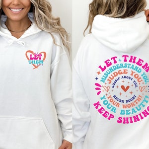 May include: White hoodie with a pink heart and the text "Let Them" on the front and a larger pink circle with the text "Let Them Misunderstand You. Judge You. Gossip About You. Never Doubt Your Worth! Keep Shining! Your Beauty!" on the back.