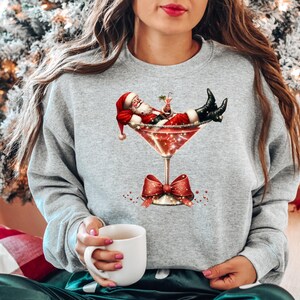 May include: A gray sweatshirt with a festive design featuring Santa Claus relaxing in a martini glass. Santa is wearing a red suit and a white beard, and he is holding a cocktail.