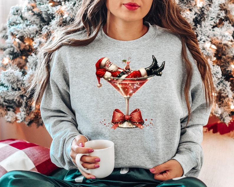 May include: A gray sweatshirt with a festive design featuring Santa Claus relaxing in a martini glass. Santa is wearing a red suit and a white beard, and he is holding a cocktail.