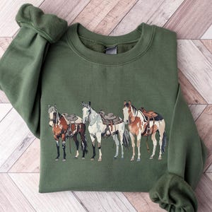May include: A green sweatshirt with a graphic of four horses wearing saddles. The horses are brown, white, and black.