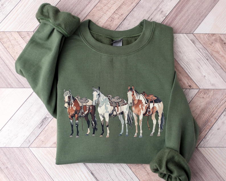 May include: A green sweatshirt with a graphic of four horses wearing saddles. The horses are brown, white, and black.