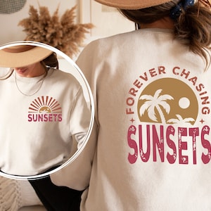 May include: White sweatshirt with a graphic print on the front and back. The front graphic says "SUNSETS" with a stylized sunset design. The back graphic says "FOREVER CHASING SUNSETS" with a palm tree design.