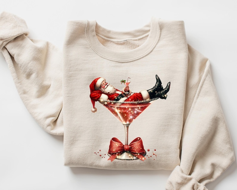 May include: A beige sweatshirt with a festive design of Santa Claus relaxing in a martini glass with a red bow.