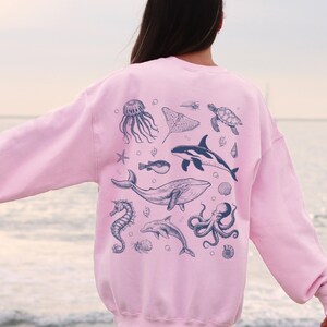 May include: A light pink sweatshirt featuring a collection of marine life illustrations in dark blue. The design includes a jellyfish, stingray, turtle, orca, whale, seahorse, dolphin, and octopus. The sweatshirt is a comfortable, casual style.