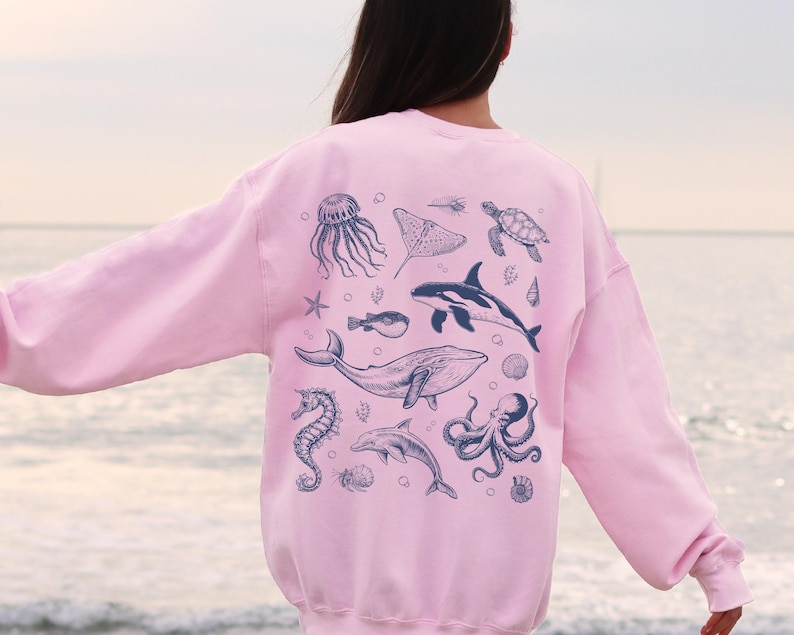 May include: A light pink sweatshirt featuring a collection of marine life illustrations in dark blue. The design includes a jellyfish, stingray, turtle, orca, whale, seahorse, dolphin, and octopus. The sweatshirt is a comfortable, casual style.