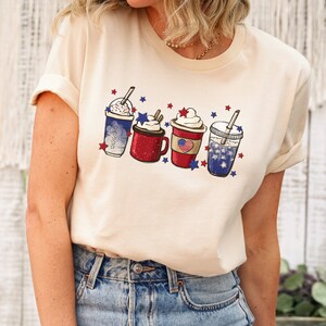 May include: A cream-colored t-shirt featuring a graphic of four festive beverages. Each drink is decorated with red, white, and blue stars and stripes, evoking a patriotic theme. The drinks include whipped cream and straws.