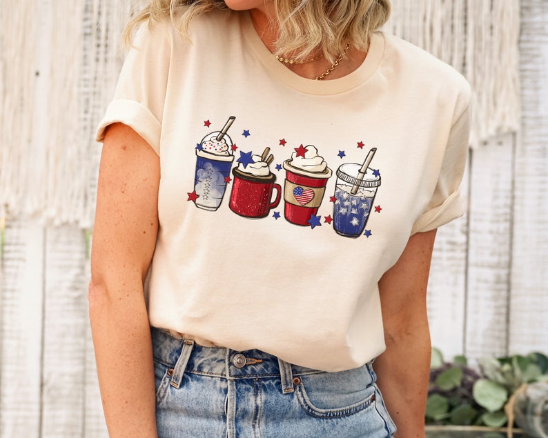 May include: A cream-colored t-shirt featuring a graphic of four festive beverages. Each drink is decorated with red, white, and blue stars and stripes, evoking a patriotic theme. The drinks include whipped cream and straws.
