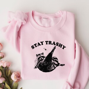 Op de afbeelding: Roze trui met een zwarte afbeelding van een wasbeer en de tekst "STAY TRASHY".