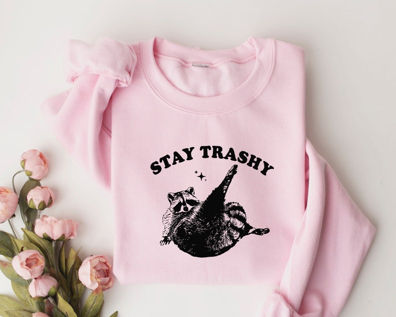 Op de afbeelding: Roze trui met een zwarte afbeelding van een wasbeer en de tekst "STAY TRASHY".